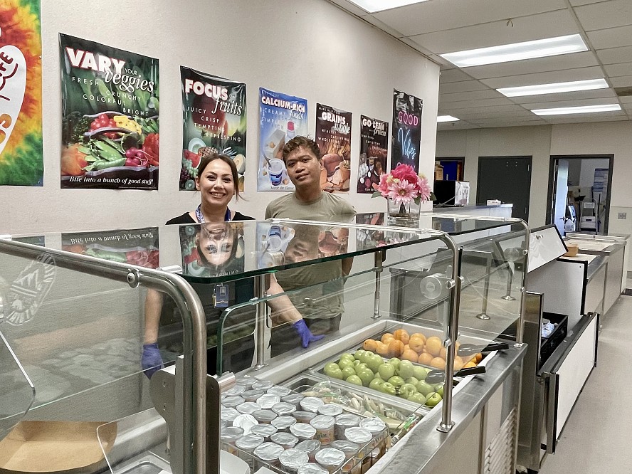 man and woman stand behind cases of fresh food in school lunch line